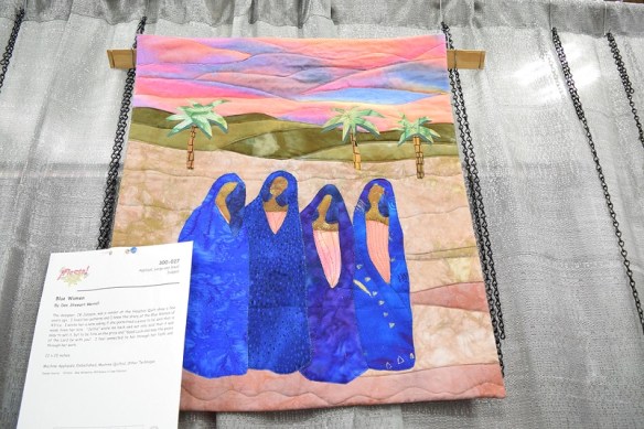 An art quilt created by Dee Merrill of four women in blue robes walking in the desert.