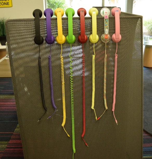 Eight phone handsets each a different color hanging on the side of a short cubicle wall.