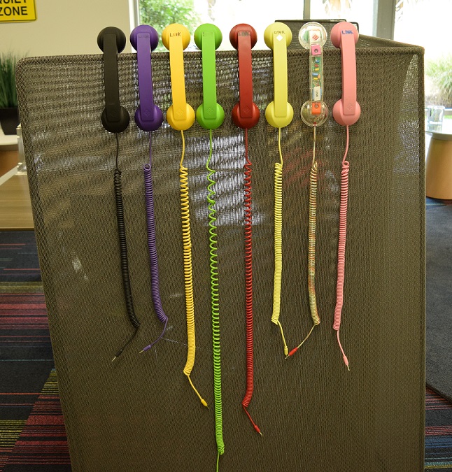 Eight phone handsets each a different color hanging on the side of a short cubicle wall.