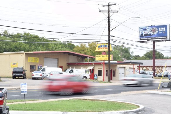 A blurred red vehicle driving by a yellow Auto Works sign and the sign is in focus. 