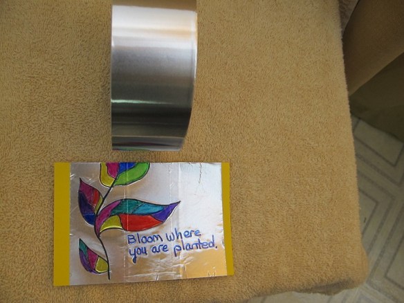 A post card decorated with foil duct tape and a mullti-colored flower and the words: Bloom Where You Are Planted