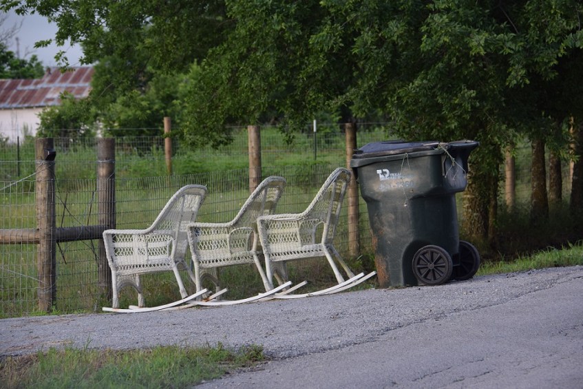 Three white wicker rocking chairs next to the trash container, to be thrown away